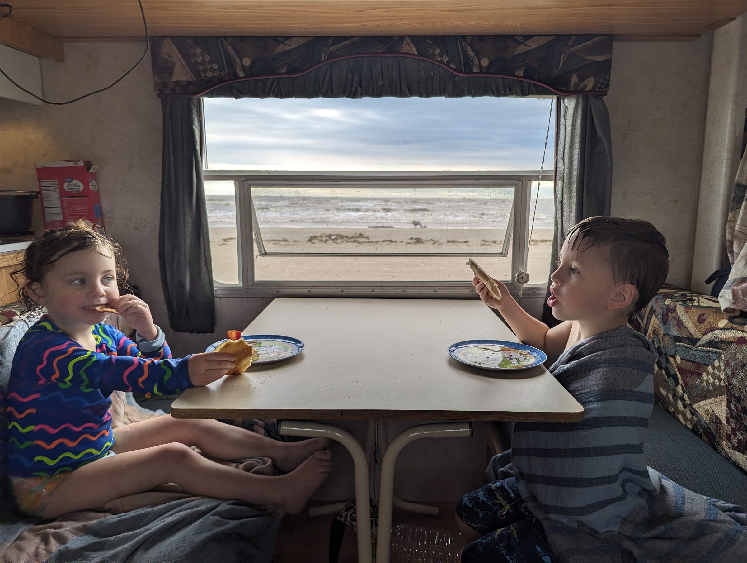 Two kids at the camper table with a beach outside the window